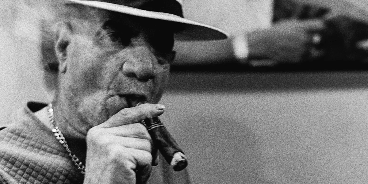This is a black and white image with a latin man wearing a panama stytle hat smoking a thick cigar the image is cropped showing his shoulders and face. His hand is also seen holding the cigar. Trees are in the background