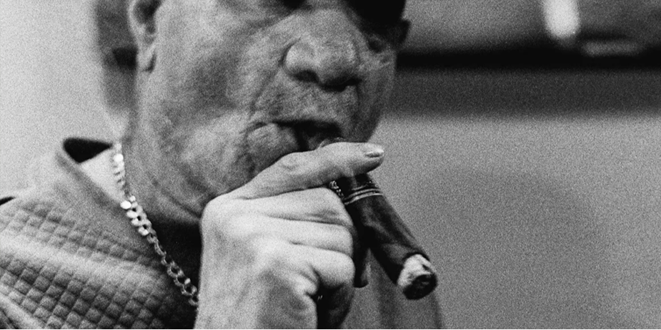 This is a black and white image with a latin man wearing a panama stytle hat smoking a thick cigar the image is cropped showing his shoulders and face. His hand is also seen holding the cigar. Trees are in the background