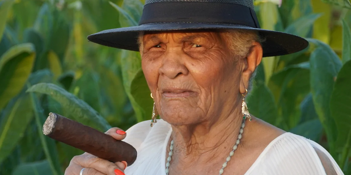 This image is of a woman standing in front of large bushes with large green leaves wearing a black hat smoking a large cigar.
