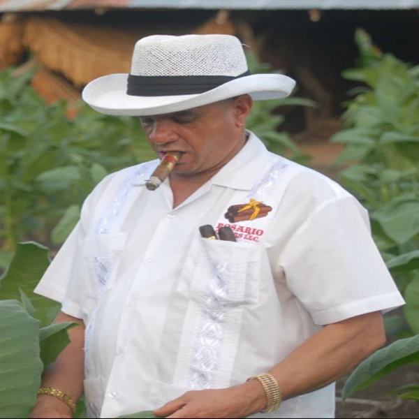a latin man dressed in white with a cigar in his mount wearing a white short sleeve shirt and hat smoking a cigar in front of large green plants