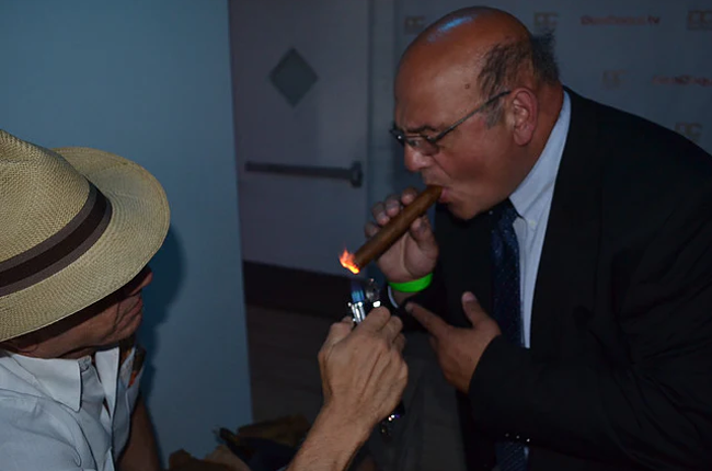This images shows a mant lighting a cigar for another men dressed in a suit with glasses on.