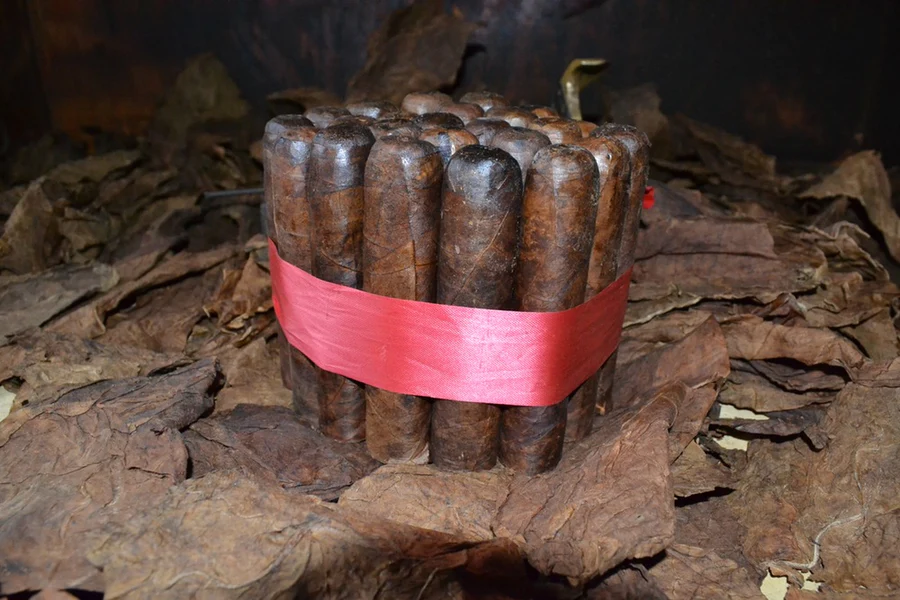 This is a image of a stack of 20 to 25 cigars. They are Rapped together with a thin colored paper ranging from cool whites to firey red wrappings they are forming a square shape simular which resembles a cube. While the Background shows the cigars placed on a feild of tabaco.