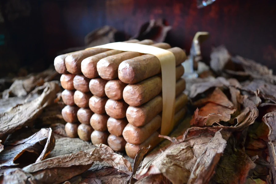 This is a image of a stack of 20 to 25 cigars. They are Rapped together with a thin colored paper ranging from cool whites to firey red wrappings they are forming a square shape simular which resembles a cube. While the Background shows the cigars placed on a feild of tabaco.