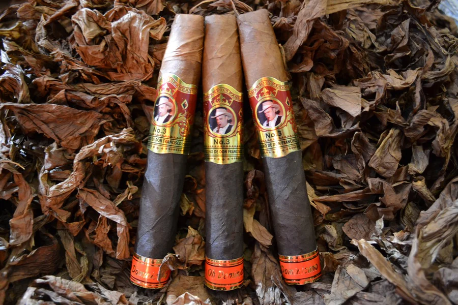 This is a image of a stack of 1 to 5 cigars. They are layed on a of Background feild of tabaco. These are two toned cigars milk chocolate and half dark chocolate in color wiht a gold labeled wrapper in the middle and a red label at the tip.