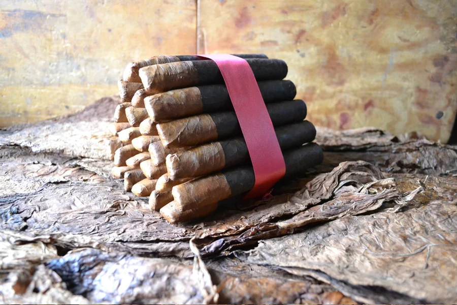 This is a image of a stack of 20 to 25 two tone cigars the top half is a lighter shade than the bottom half. They are Rapped together with a thin colored paper ranging from cool whites to firey red wrappings they are forming a trianglur shape simular to a mountain or pyramid. While the Background shows the cigars placed on a feild of tabaco.