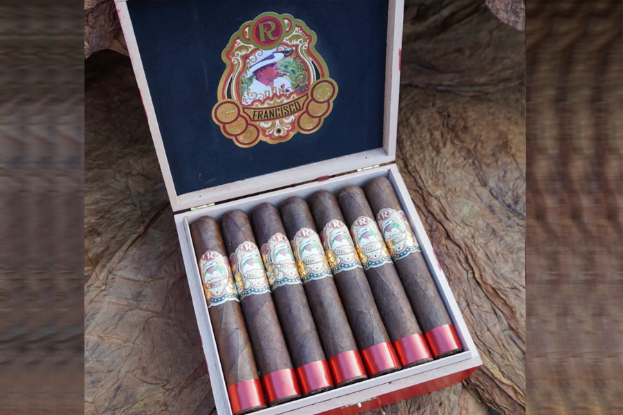 This is a image of a stack of 7 or more cigars. They are Pleaced together in a small wooden box that has a cover to it with the company's logo. This image is zoomed out to show the image at a distance.