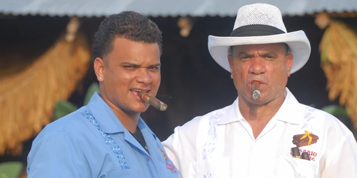 This image is of two men standing together smoking cigars. on is younger with a blue button up shirt and the other is older with a white shirt and hat.