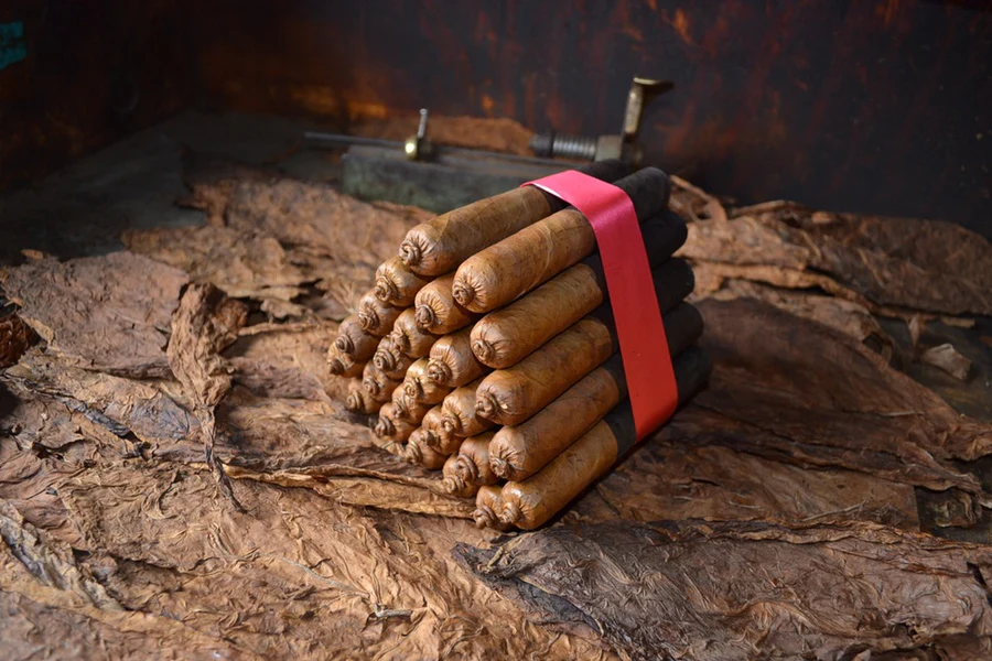 This is a image of a stack of 20 to 25 cigars. They are Rapped together with a thin colored paper ranging from cool whites to firey red wrappings they are forming a trianglur shape simular to a mountain or pyramid. While the Background shows the cigars placed on a feild of tabaco.