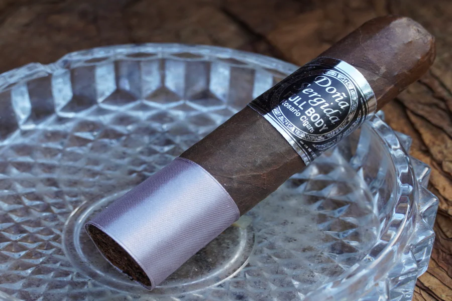 This imade is of a dark colored stubby cigar in a clean round crystal ashtray. The cigar is decorated with a silver rapper in the mid sectoin as well as the bottom quarter.
