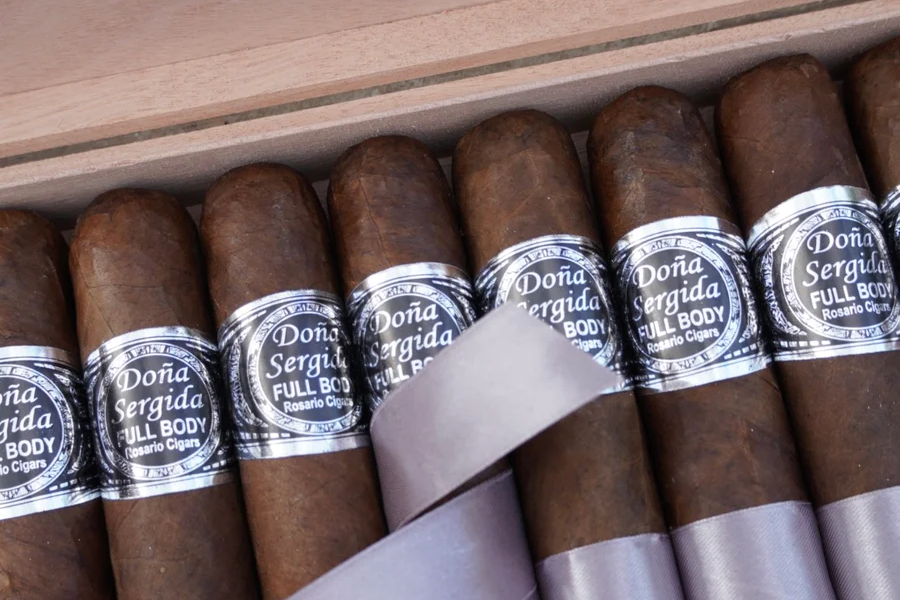 This is a image of a stack of 10 or more cigars. They are Pleaced together in a small wooden box that has a cover to it with the company's logo that is wrapped in a circular reef like design. This is a close up of the first image of the same product.