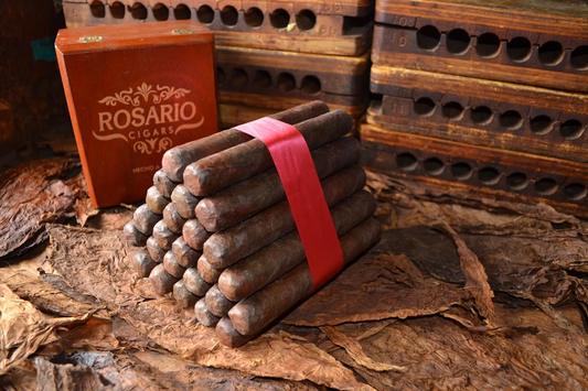 This is a image of a stack of 20 to 25 cigars. They are Rapped together with a thin colored paper ranging from cool whites to firey red wrappings they are forming a trianglur shape simular to a mountain or pyramid. While the Background shows the cigars placed on a feild of tabaco. there is also a red box container in the background that says Rosario Cigars written on it.