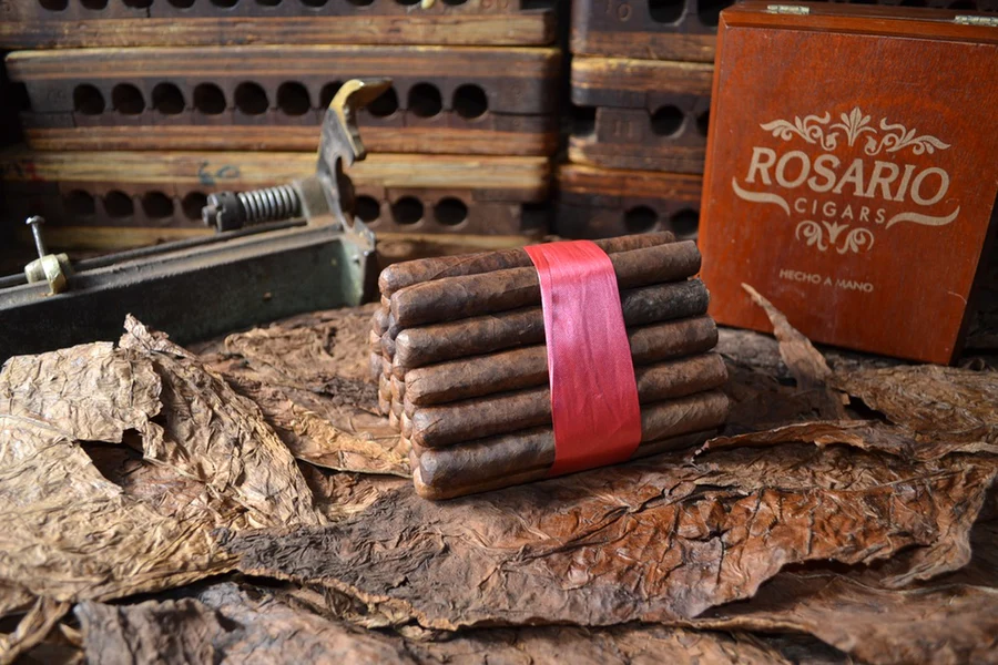This is a image of a stack of 20 to 25 cigars. They are Rapped together with a thin colored paper ranging from cool whites to firey red wrappings they are forming a trianglur shape simular to a mountain or pyramid. While the Background shows the cigars placed on a feild of tabaco. There is also a retangular case in tgeh backgound that has the name Rosario Cigars written on it.