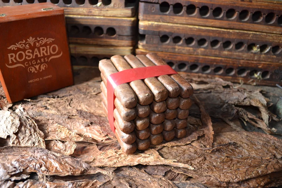 This is a image of a stack of 20 to 25 cigars. They are Rapped together with a thin colored paper ranging from cool whites to firey red wrappings they are forming a trianglur shape simular to a mountain or pyramid. While the Background shows the cigars placed on a feild of tabaco. There is alkso a red retangular case in the background that says Rosario Cigars onit.