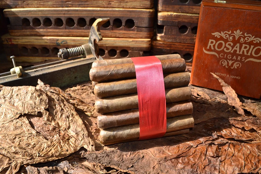 This is a image of a stack of 20 to 25 cigars. They are Rapped together with a thin colored paper ranging from cool whites to firey red wrappings they are forming a trianglur shape simular to a mountain or pyramid. While the Background shows the cigars placed on a feild of tabaco. There is also a red retangular case in the background that has the words Rosario Cigars written on it.