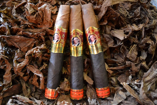 This is a image of a stack of 1 to 5 cigars. They are layed on a of  Background feild of tabaco. These are two toned cigars milk chocolate and half dark chocolate in color wiht a gold labeled wrapper in the middle and a red label at the tip.