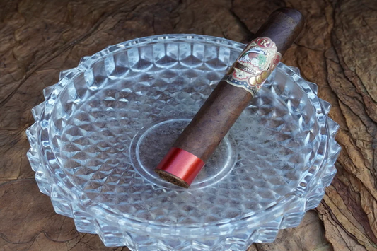 This imade is of a dark colored stubby cigar in a clean round crystal ashtray. The cigar is decorated with a gold rapper in the mid sectoin as well as red at the bottom quarter.